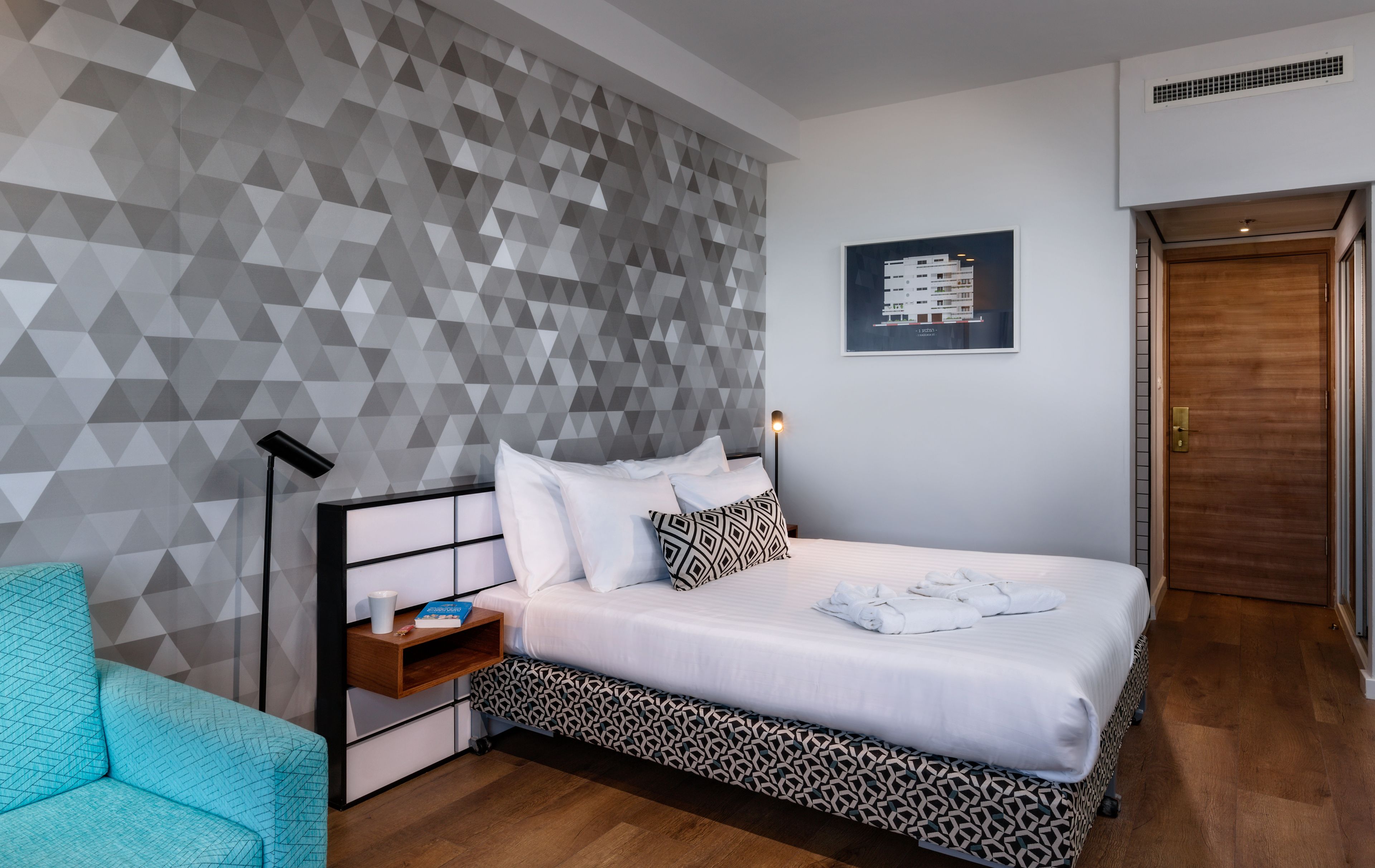 Quarto de hotel moderno com parede de padrão geométrico, cama bem arrumada com lençóis brancos, mesa de cabeceira com abajur e poltrona azul.