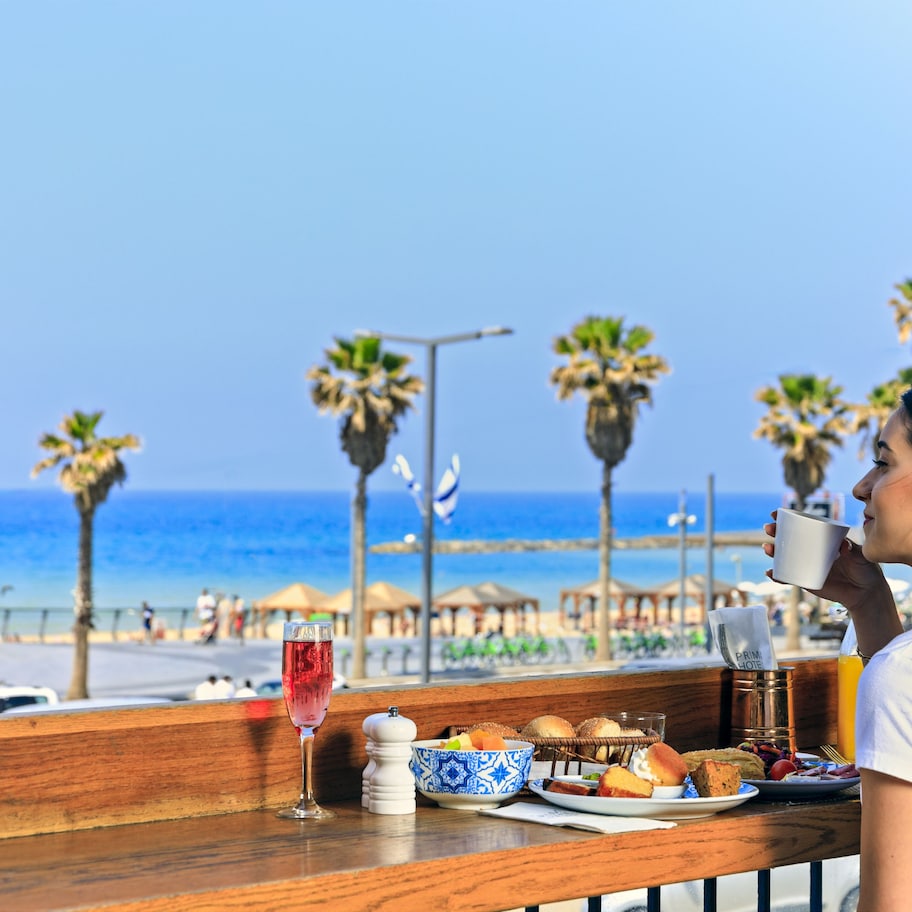 A woman sitting at a table with a view of the beach, a drink, and a plate of food.