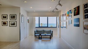 Una galería amplia con diseño moderno, que presenta un área de estar central, ventanas grandes con vista al mar y diversas obras de arte expuestas en las paredes.