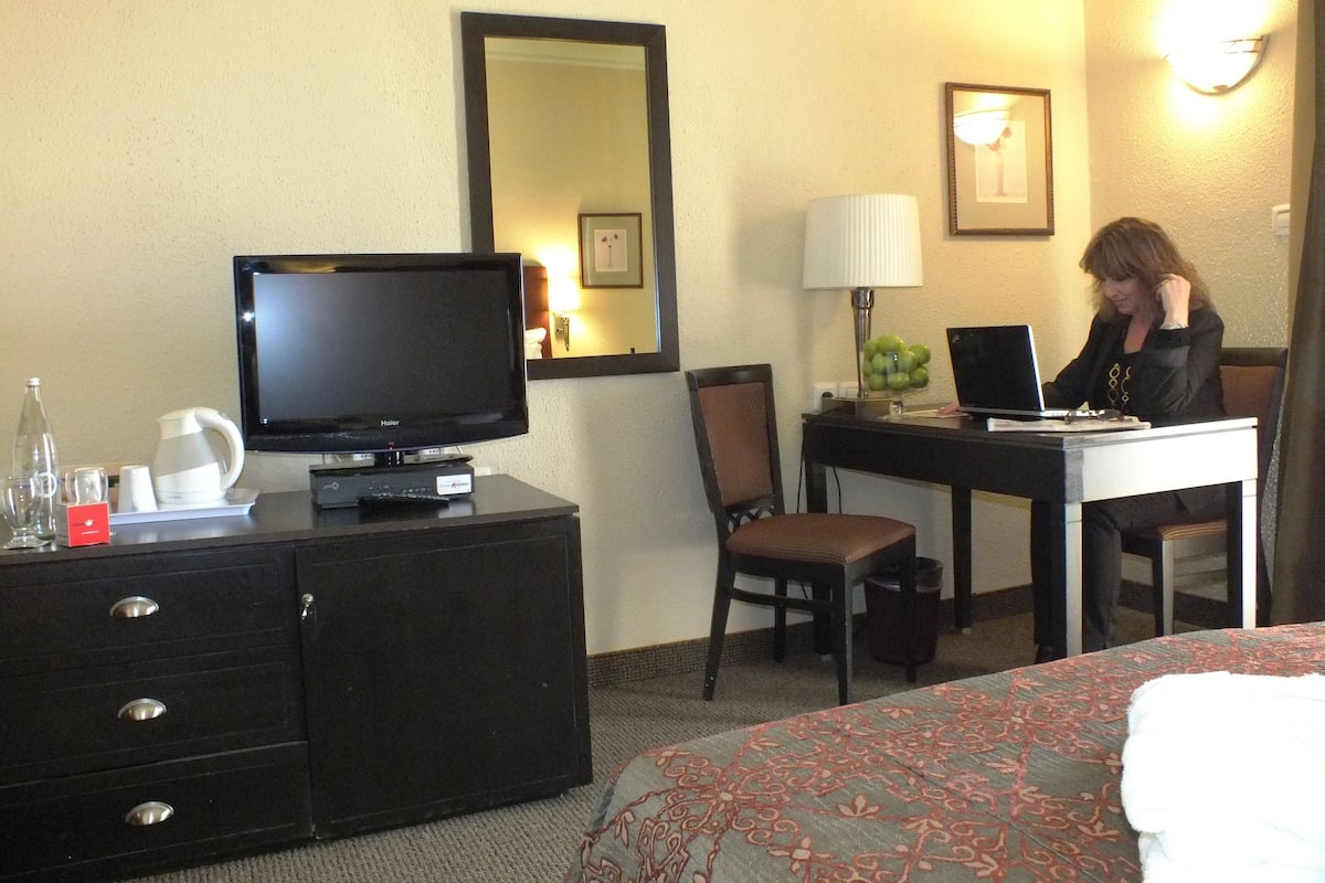A hotel room with a television, a desk with a laptop, a chair, a bed with a patterned bedspread, a dresser, and a mirror on the wall.