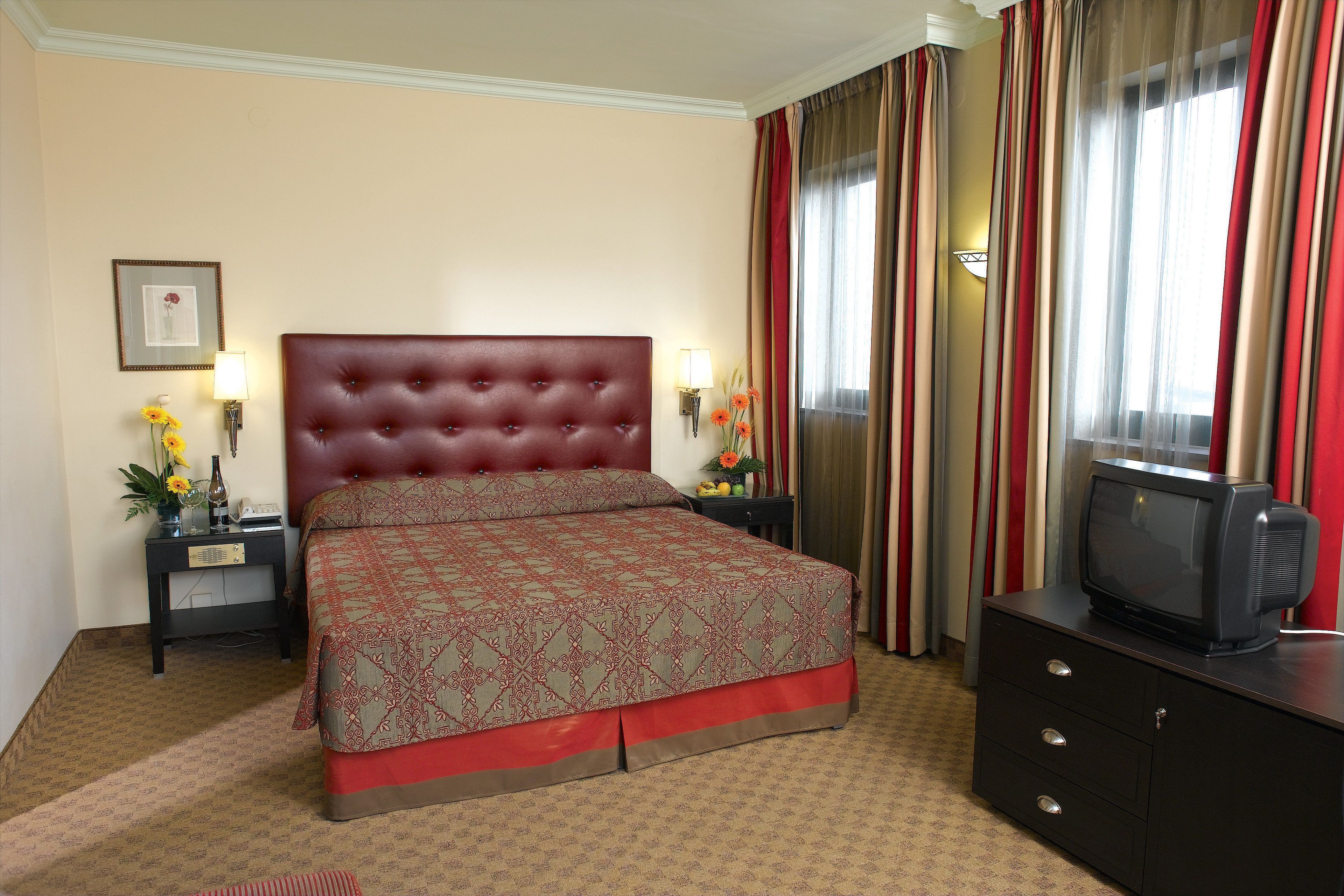 Una habitación de hotel con una cama grande, un televisor sobre una cómoda y una mesita de noche con una lámpara y flores.