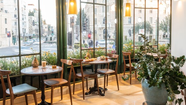 A café with round tables and chairs, large windows with a view of the street, hanging pendant lights, and a large potted plant. - Prima Kings Hotel (Jerusalem)
