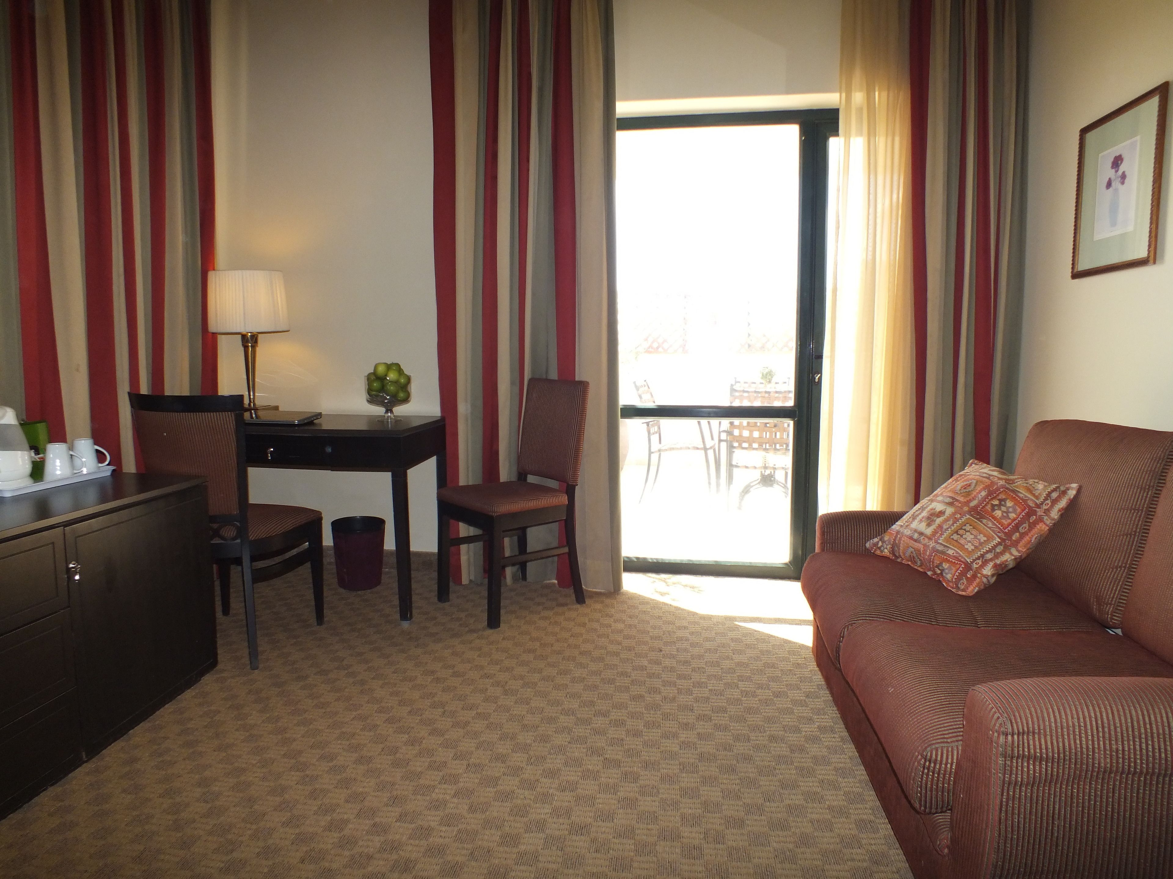A hotel room with a sofa, a desk, a chair, a lamp, and a view of the outside balcony.