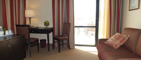 A hotel room with a sofa, a desk, a chair, a lamp, and a view of the outside balcony.