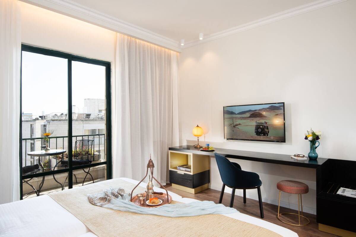 A hotel room with a large bed, a desk with a lamp, a chair, a small table with a vase, and a balcony with a view of the city.