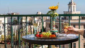 A balcony with a table set for two, featuring a fruit platter and two glasses of white wine, with a decorative vase and a view of buildings in the background. - Prima Kings Hotel (Jerusalem)