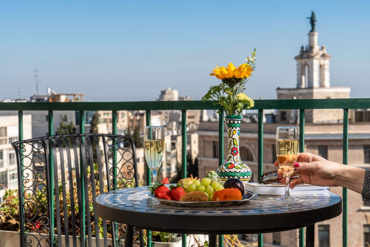A balcony with a table set for two, featuring a fruit platter and two glasses of white wine, with a decorative vase and a view of buildings in the background.