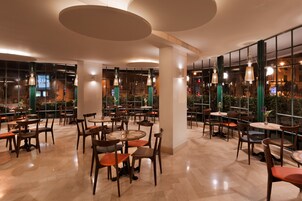 Un amplio comedor interior con varias mesas y sillas, ventanas grandes y lámparas colgantes.
