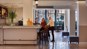 A modern bar area with a high bar counter, seating with stools, and a view of the outdoors through large windows. - Prima Kings Hotel (Jerusalem)