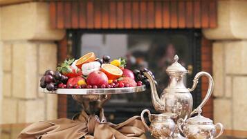 Una mesa con una bandeja de frutas, una tetera de plata y tazas, sobre un mantel frente a una chimenea.