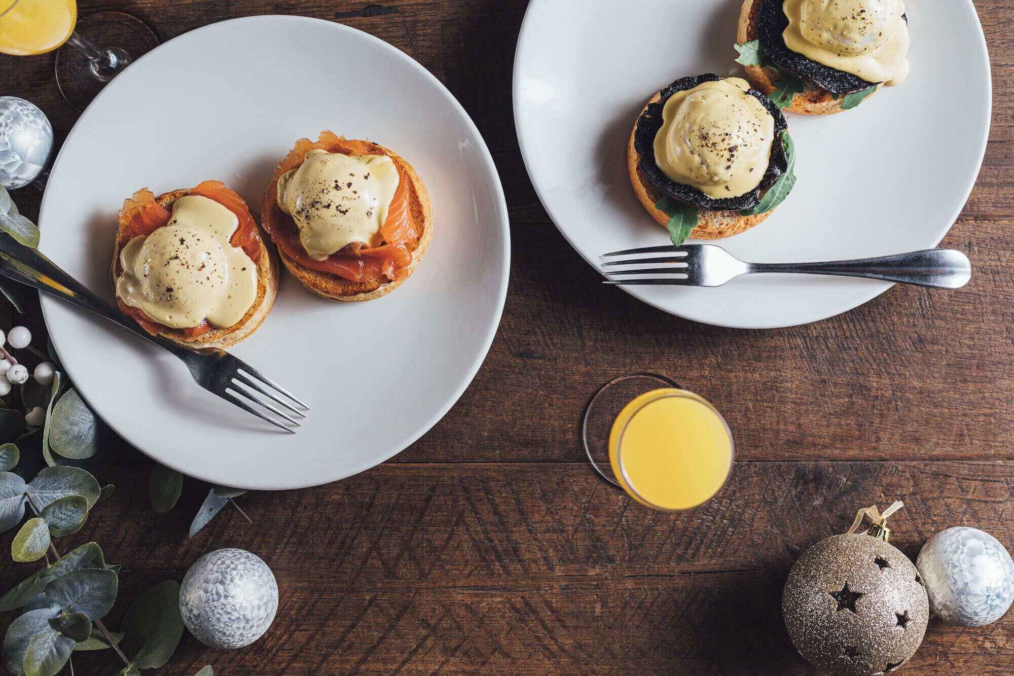 Two plates of eggs benedict with hollandaise sauce, accompanied by forks and a glass of orange juice on a wooden table.