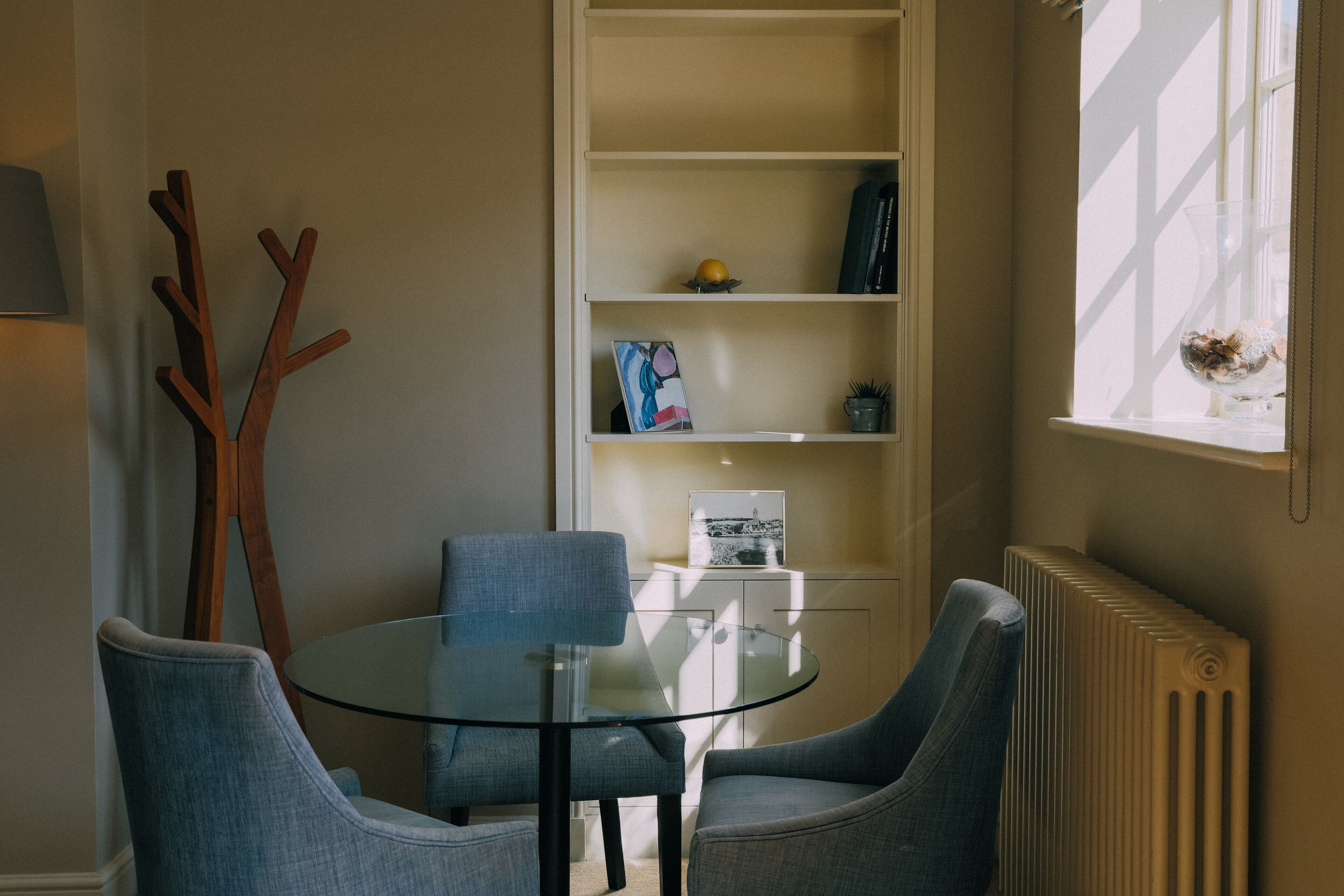 A modern room with a round glass table, two grey armchairs, a white bookshelf, and a radiator.