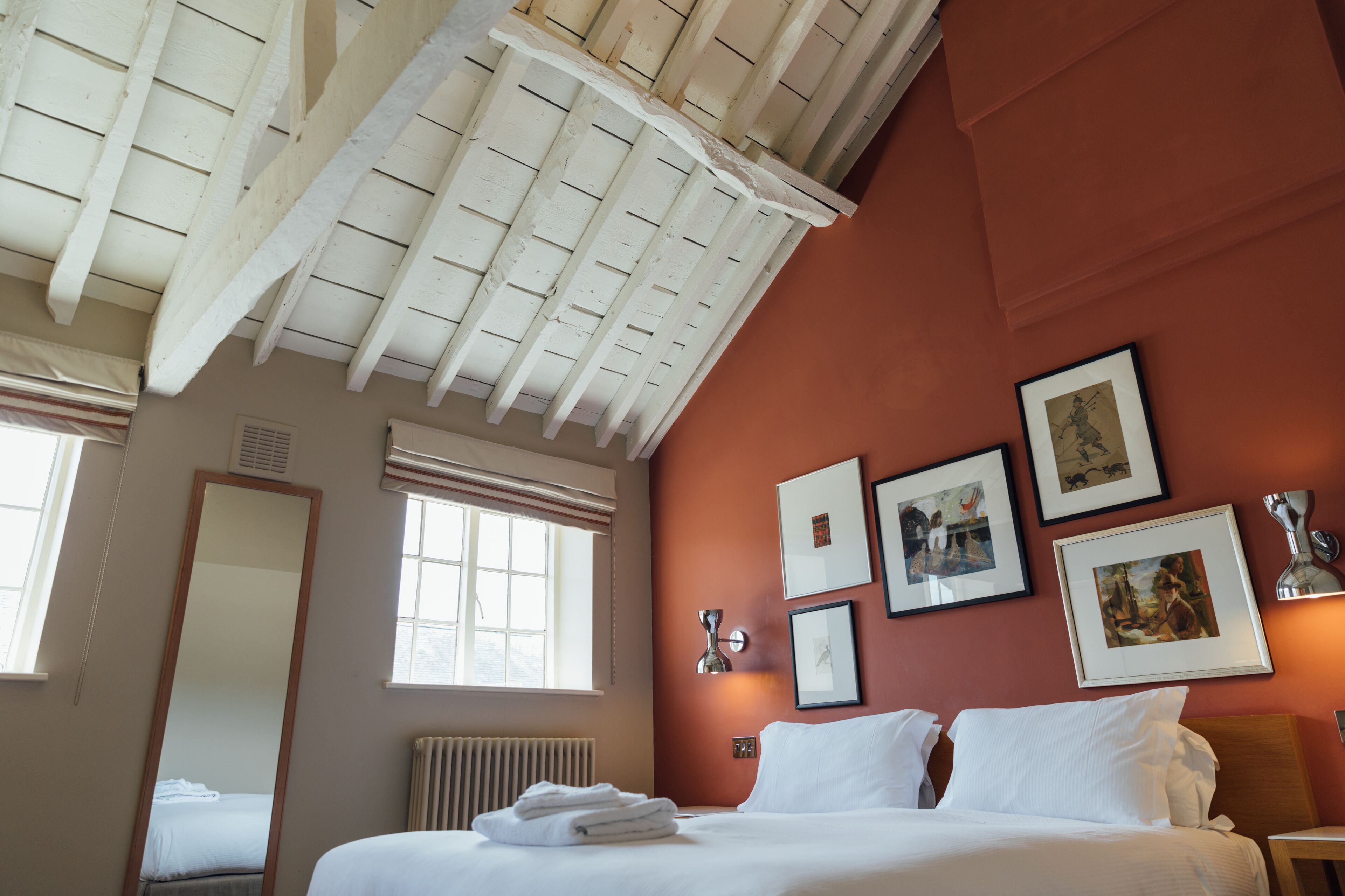 A hotel room with a large bed, a ceiling with exposed beams, a radiator, and several framed pictures on the wall.