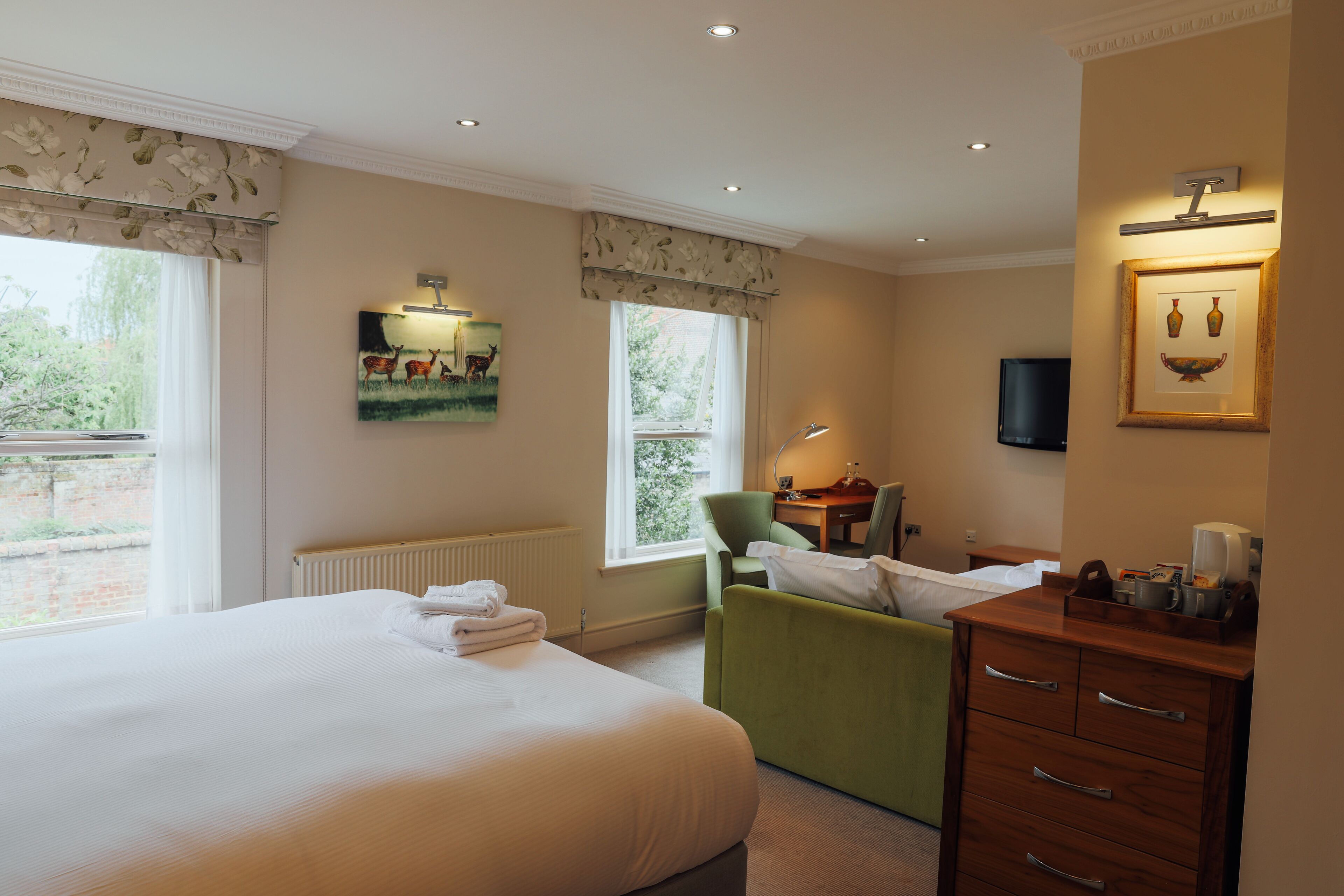 Un cuarto de hotel con una cama grande, un sillón verde, un escritorio con lámpara y un cuadro enmarcado en la pared.