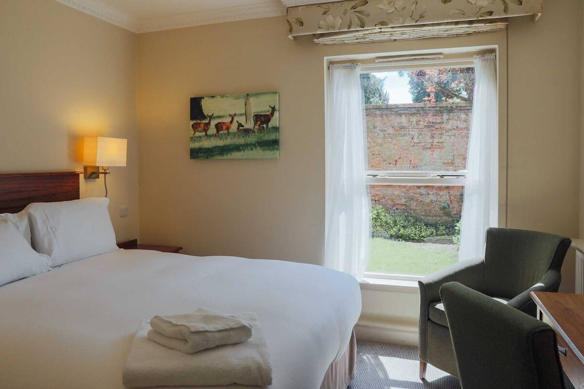 Habitación de hotel con cama, una silla, un cuadro de ciervos, ventana con vista a un muro de ladrillo y follaje, y una lámpara en la pared.