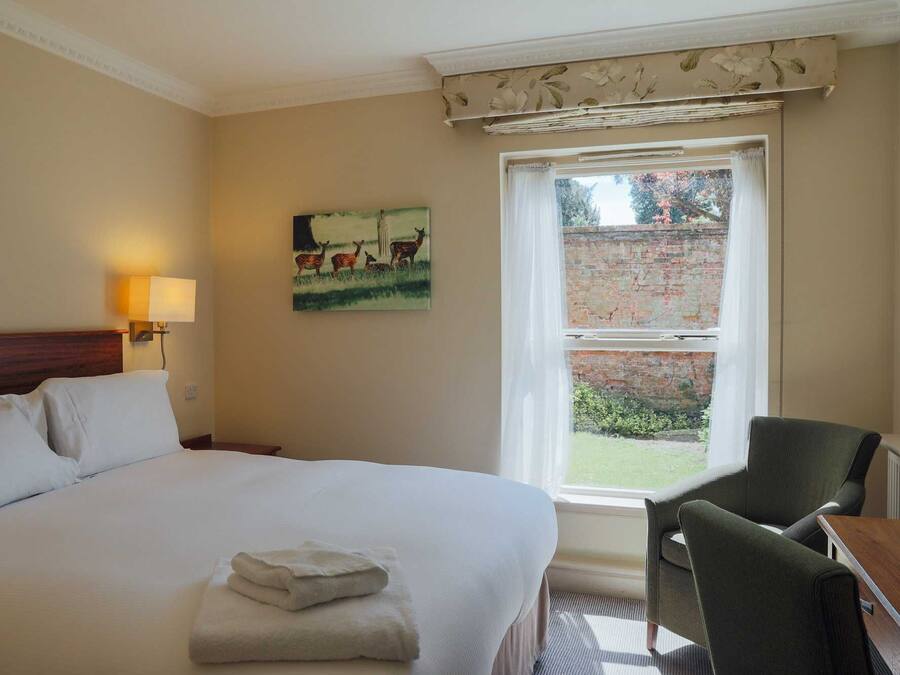Hotelkamer met een bed, een stoel, een schilderij van herten, een raam met uitzicht op een bakstenen muur en groen, en een lamp aan de muur.