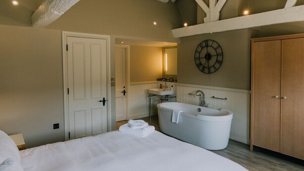 A bedroom with a large bed, a white bathtub, a wooden wardrobe, and a wall-mounted clock.