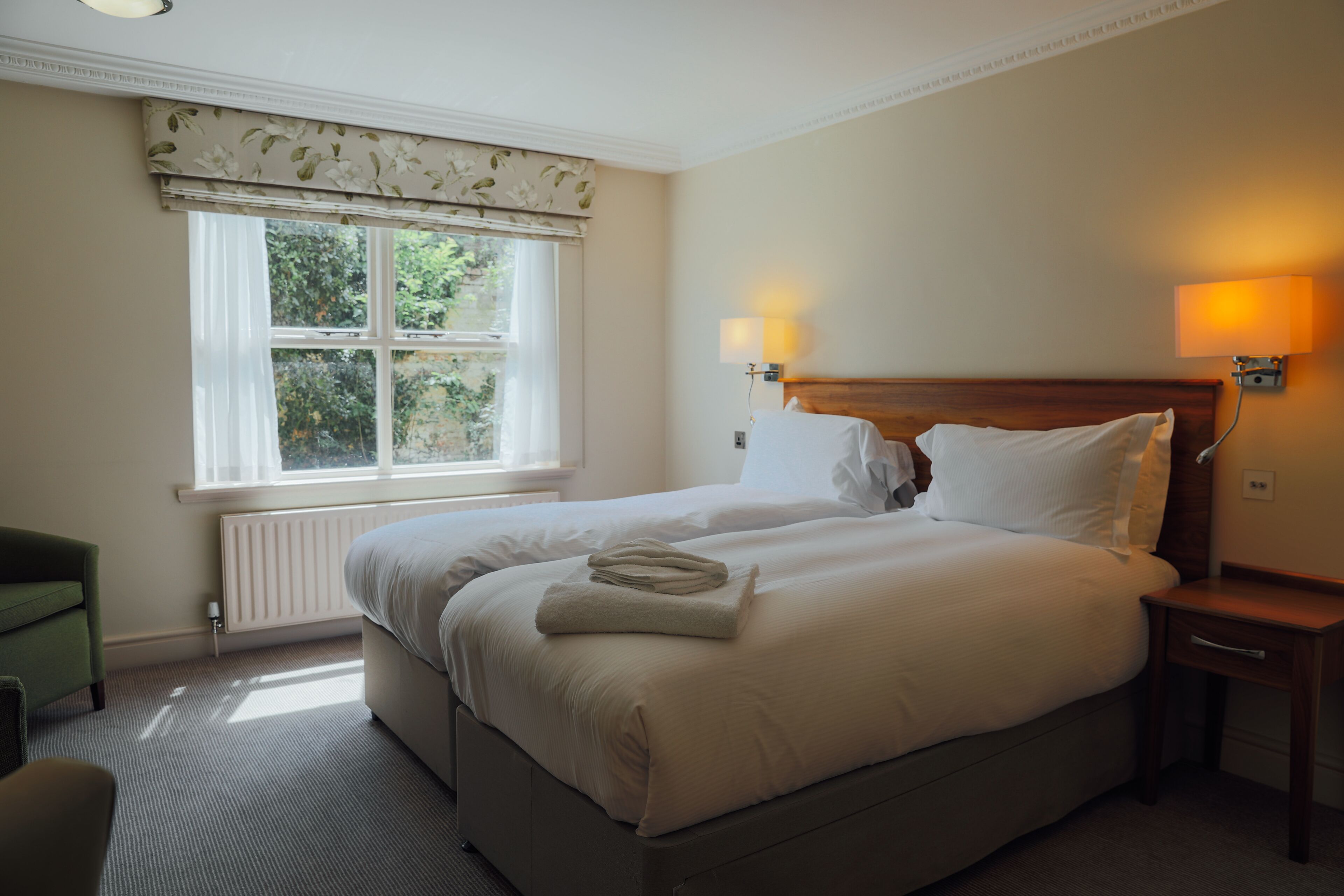 A neatly made bed with white linens, a green armchair, and a window with a view of greenery.