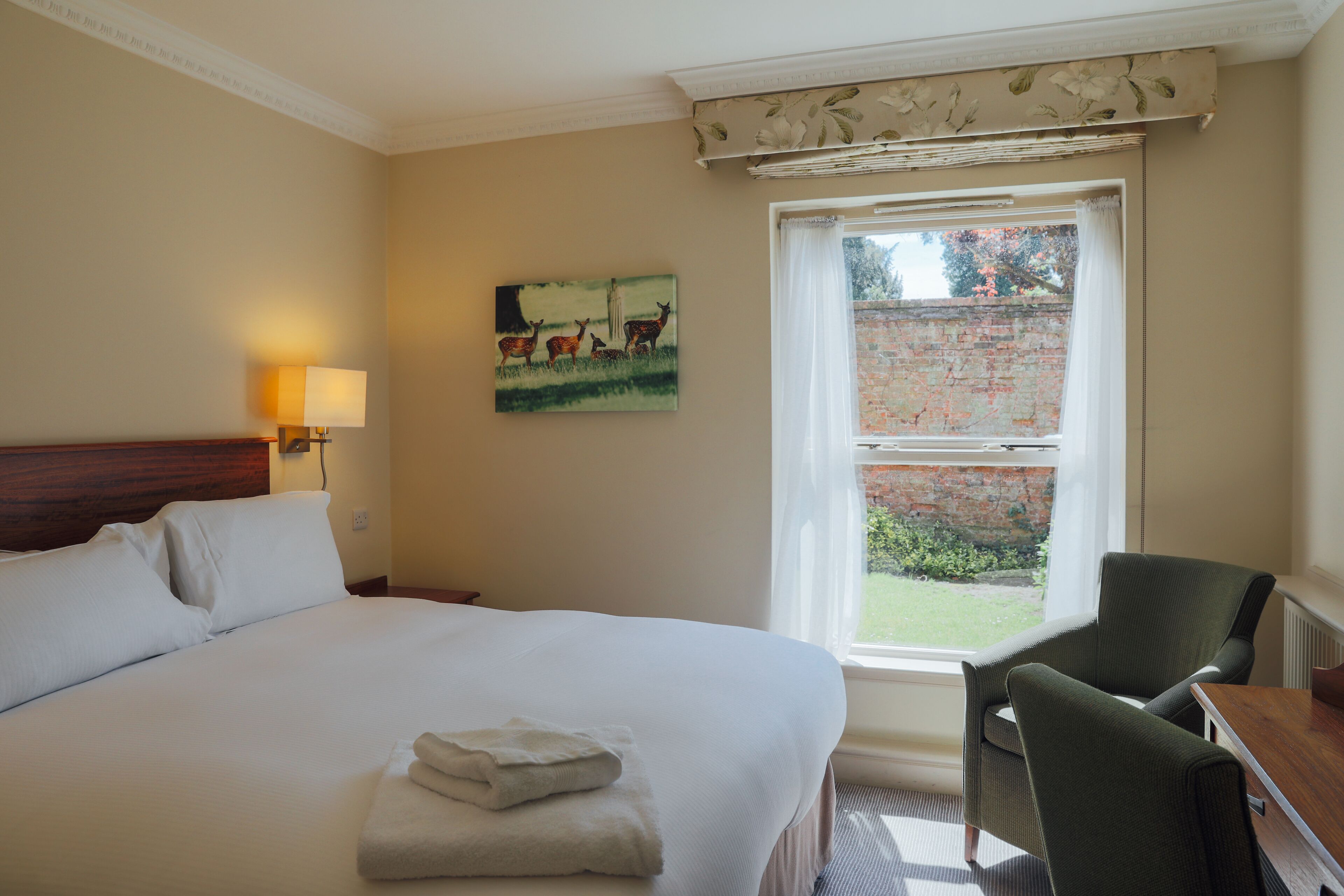 Habitación de hotel con una cama grande, un escritorio, una silla, una ventana que da a una pared de ladrillo y vegetación, y un cuadro en la pared.