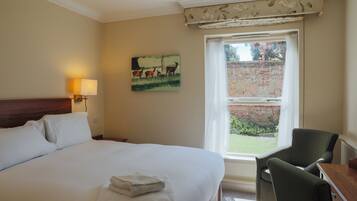 Habitación de hotel con una cama grande, un escritorio, una silla, una ventana que da a una pared de ladrillo y vegetación, y un cuadro en la pared.