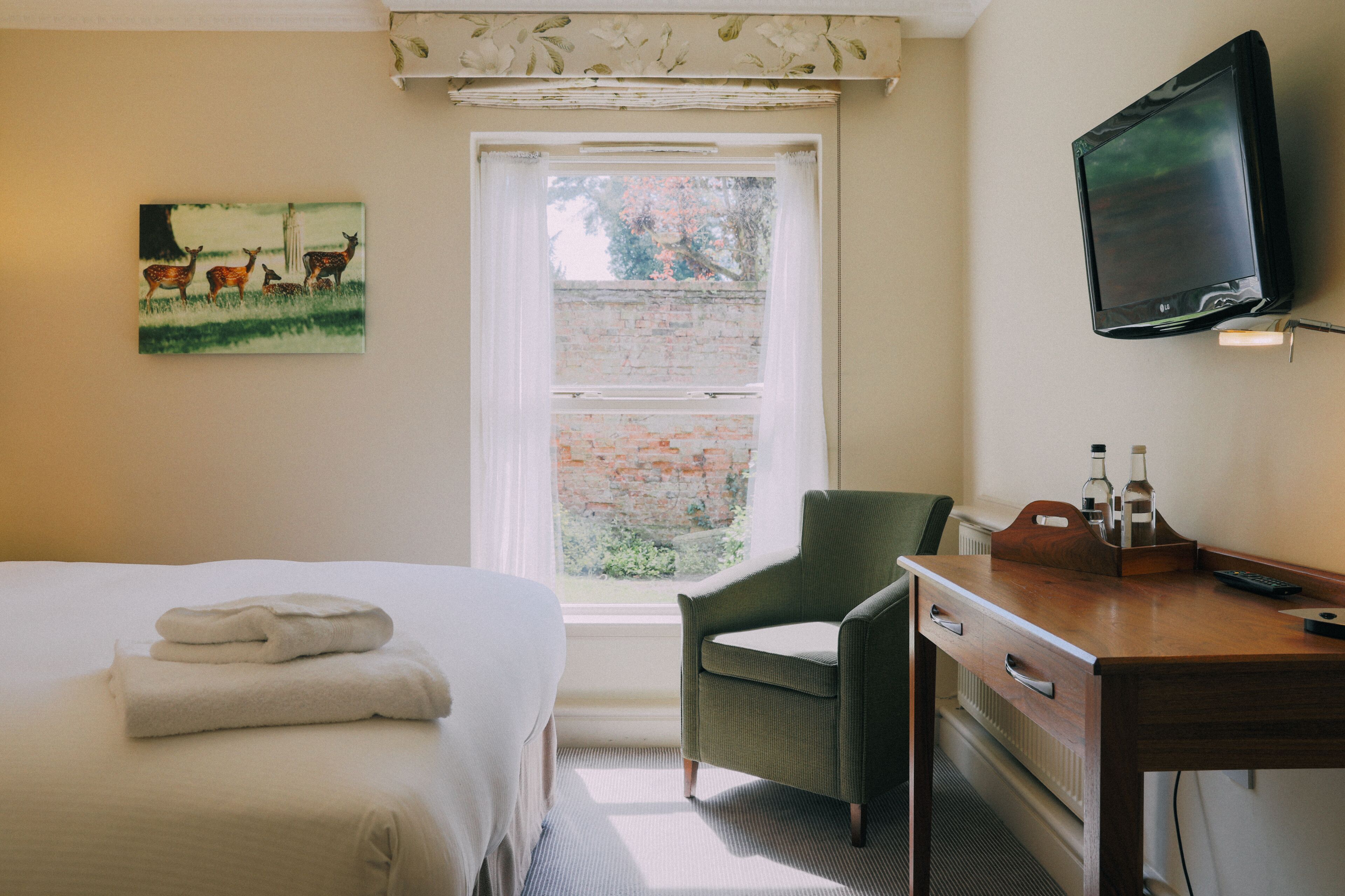 A hotel room with a bed, a chair, a desk with a television and remote, a window with a view of greenery, and a painting on the wall.