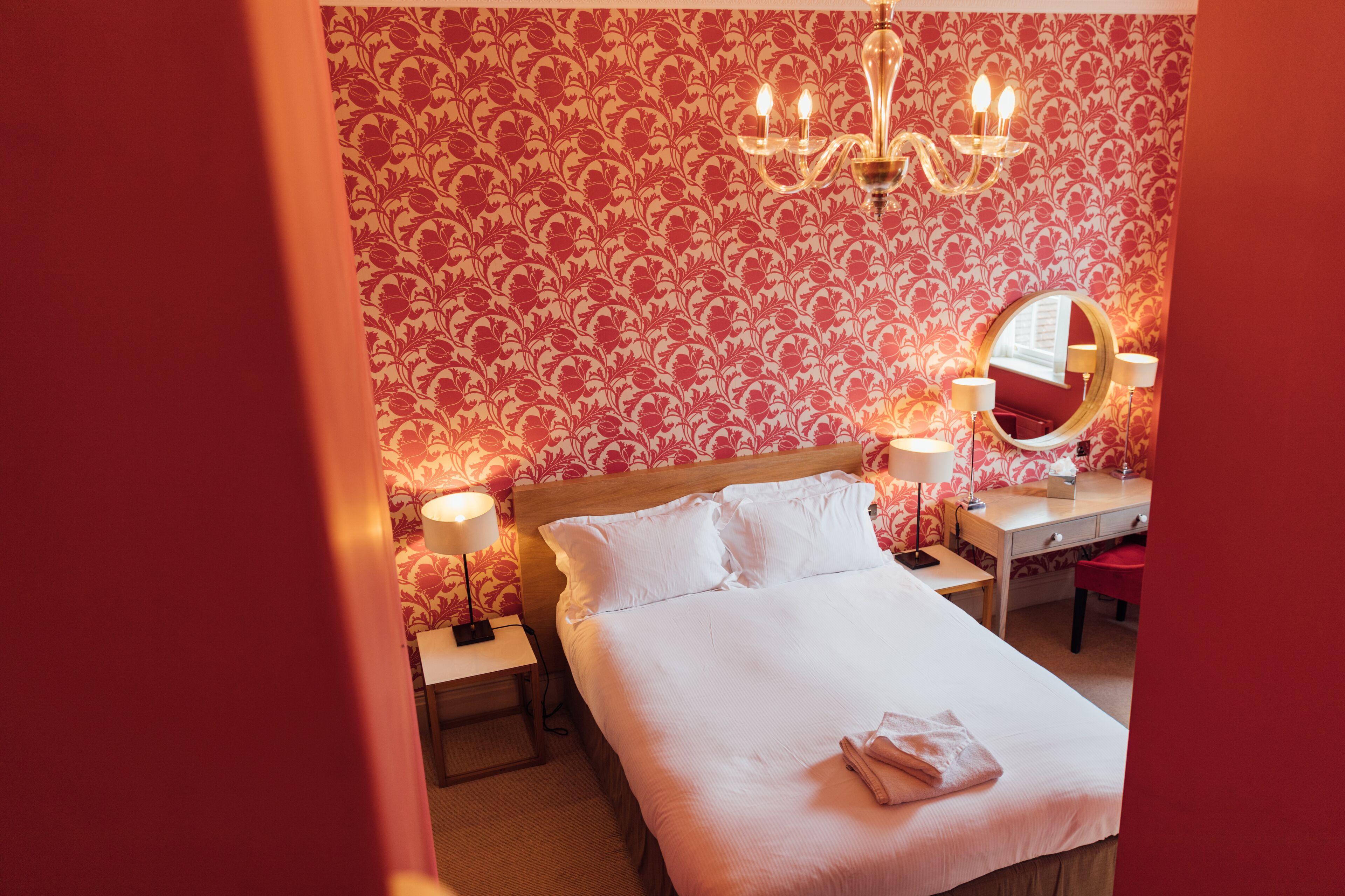 Een hotelkamer met een groot bed, twee nachtkastjes met lampen, een ronde spiegel en een kroonluchter.