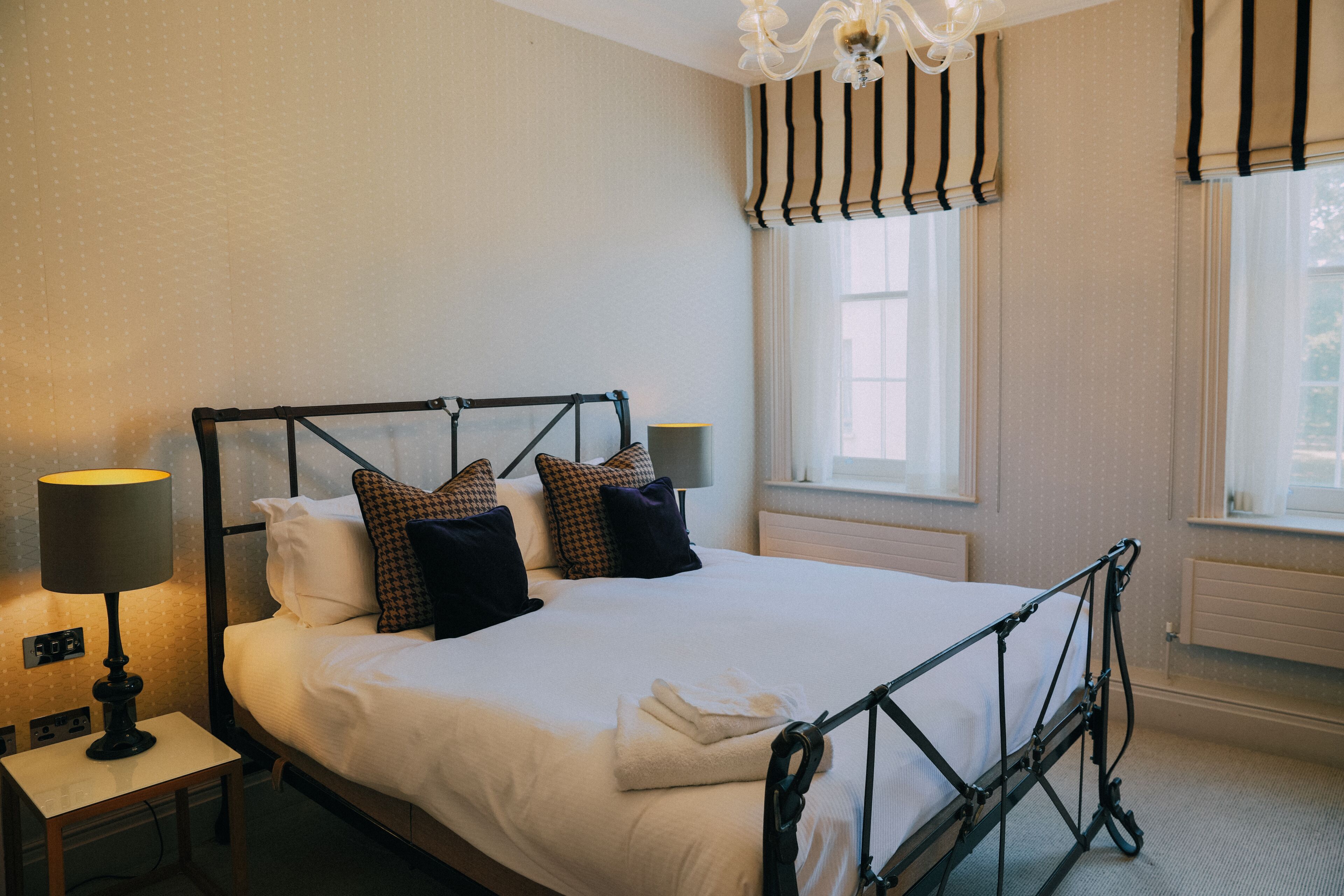 Una cama bien hecha con estructura metálica, una lámpara de noche y una ventana con cortinas.