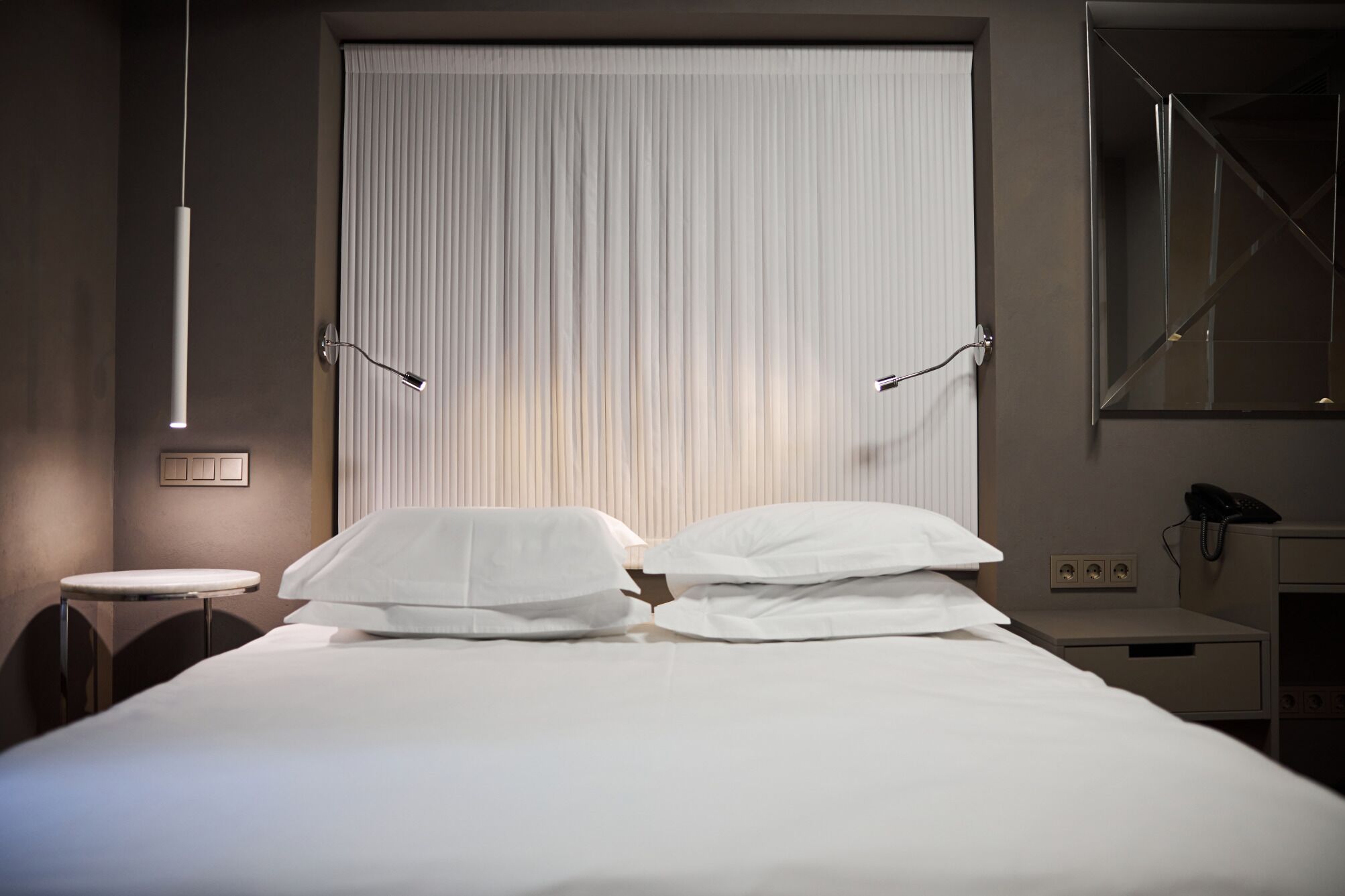 Habitación de hotel con una cama grande, una mesita de noche, un escritorio, una silla, un espejo y una ventana con persianas.