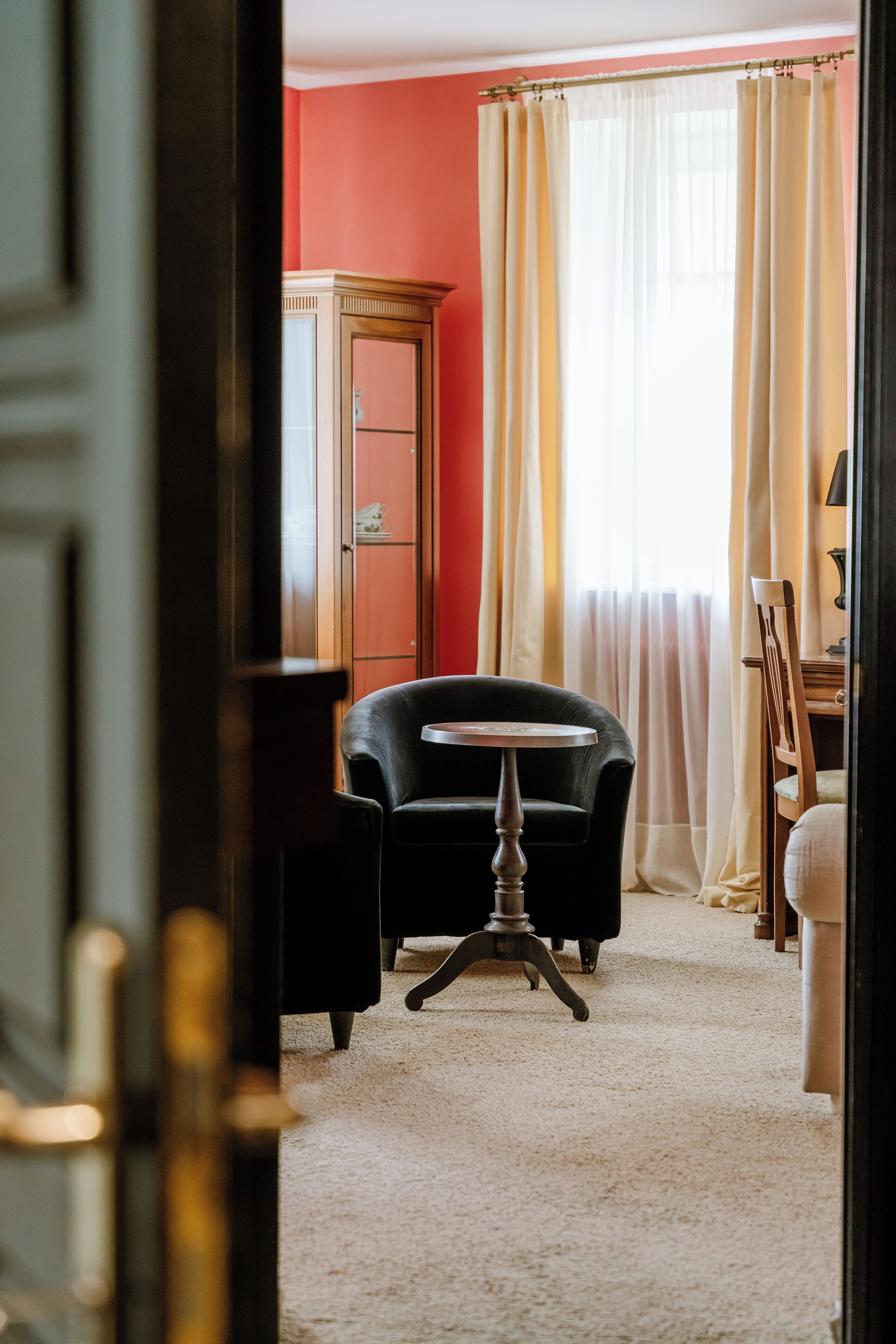 Une pièce avec un mur rouge, un fauteuil en cuir noir, une armoire en bois et une fenêtre ornée de rideaux légers.