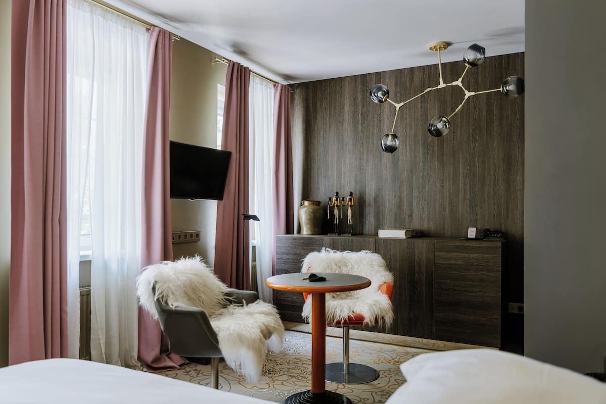 Ett hotellrum med en säng, ett runt bord med en stol, en TV, en minibar och en dekorativ taklampa.