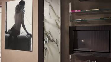 A modern bathroom with a marble shower enclosure, a mounted television, and a pink storage unit.