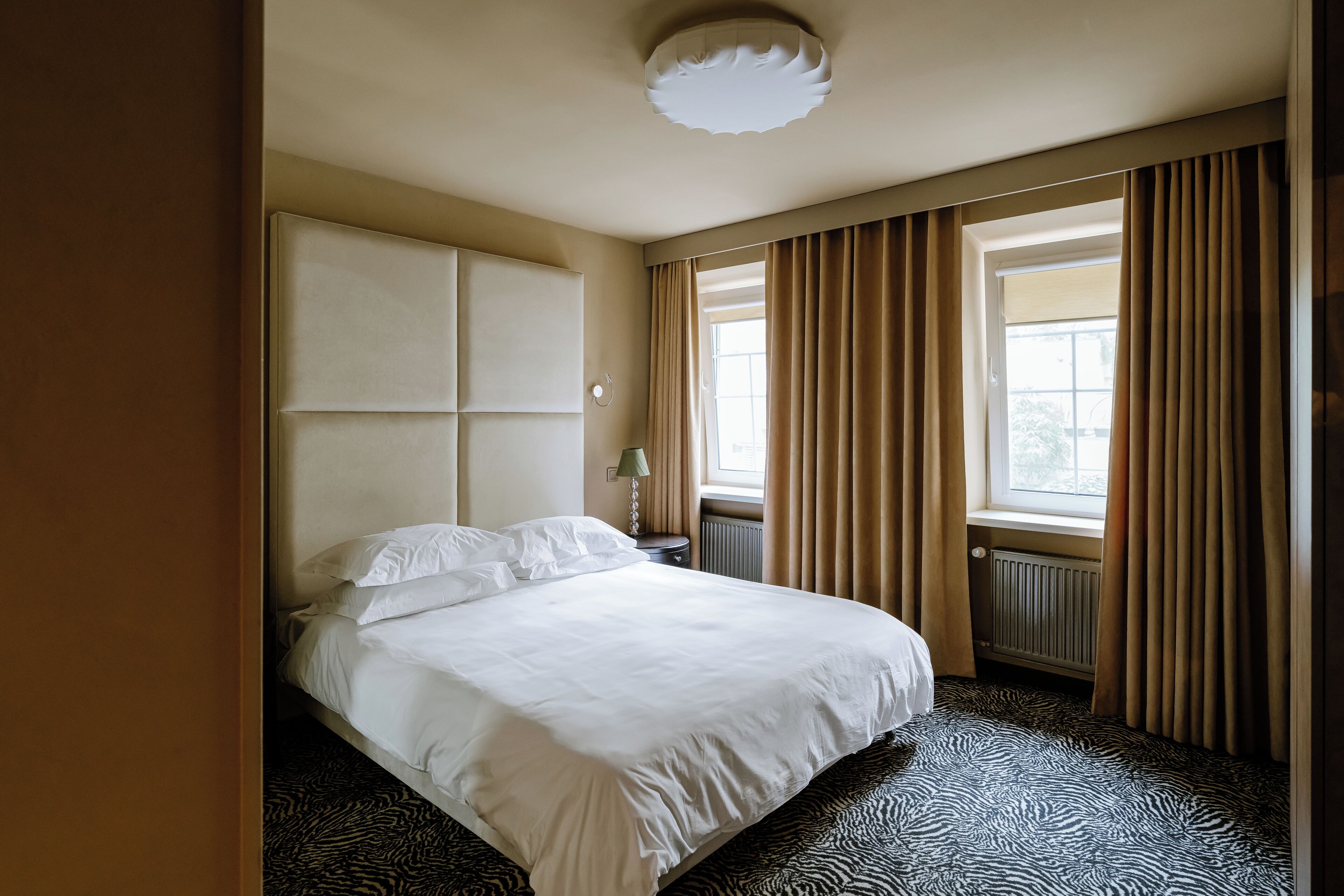 Ein Hotelzimmer mit einem großen Bett, einem Heizkörper, einem Fenster mit Vorhängen und einer Deckenleuchte.