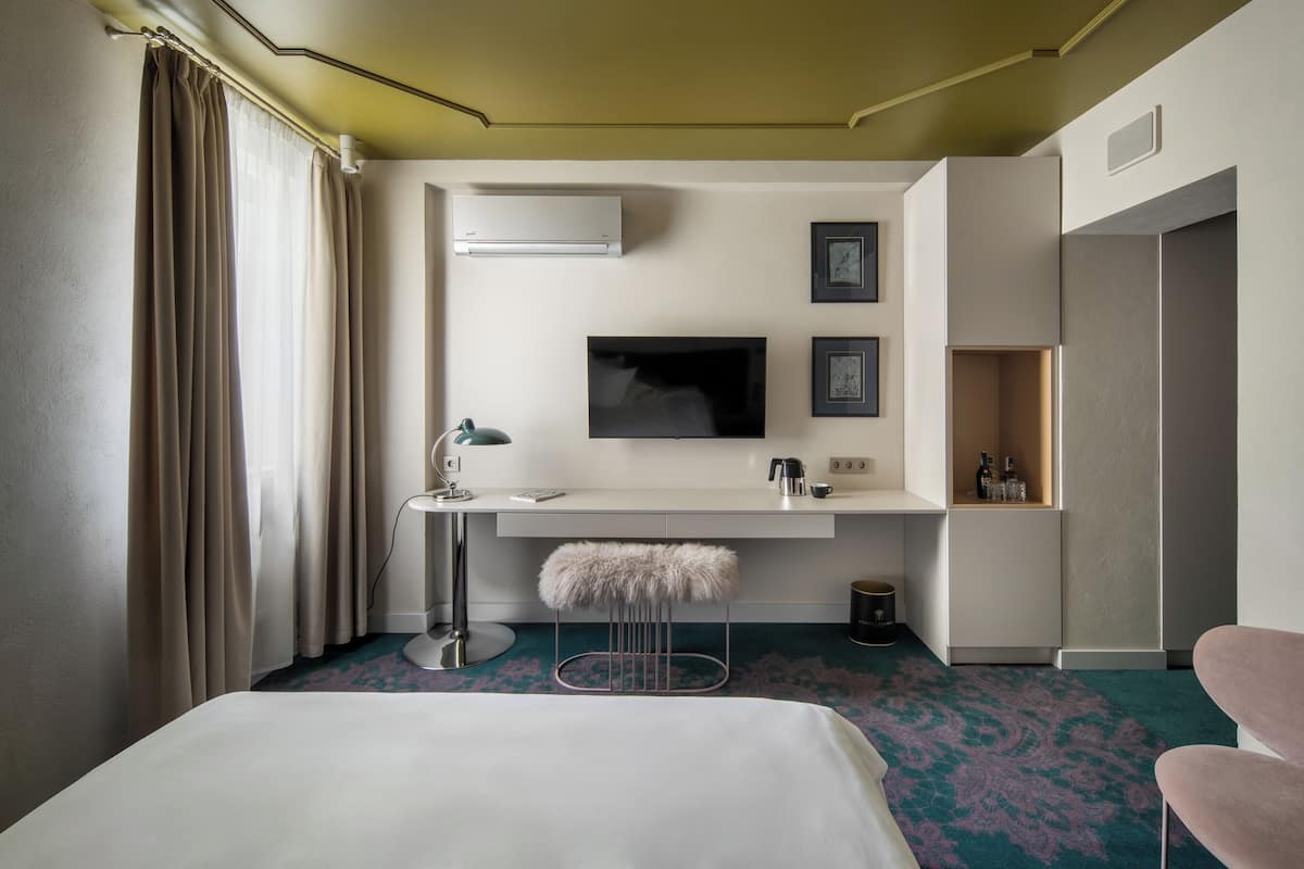 Ett hotellrum med en säng, ett skrivbord med en lampa, en tv, en minibar och en dörr till badrummet.
