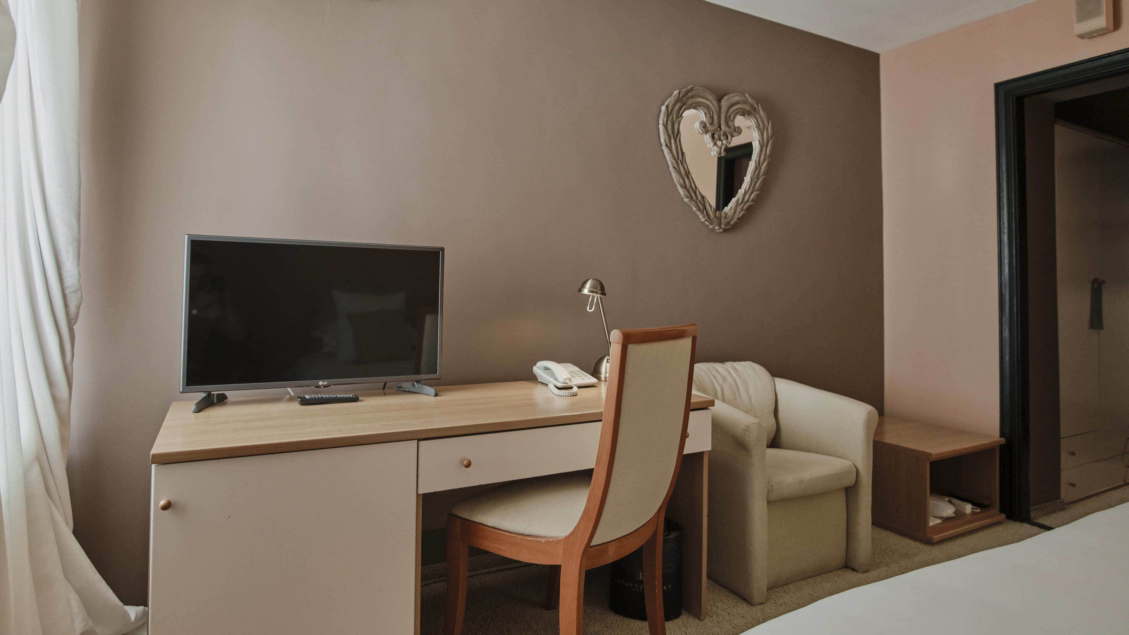 A hotel room with a desk, chair, TV, lamp, phone, and a heart-shaped mirror on the wall.