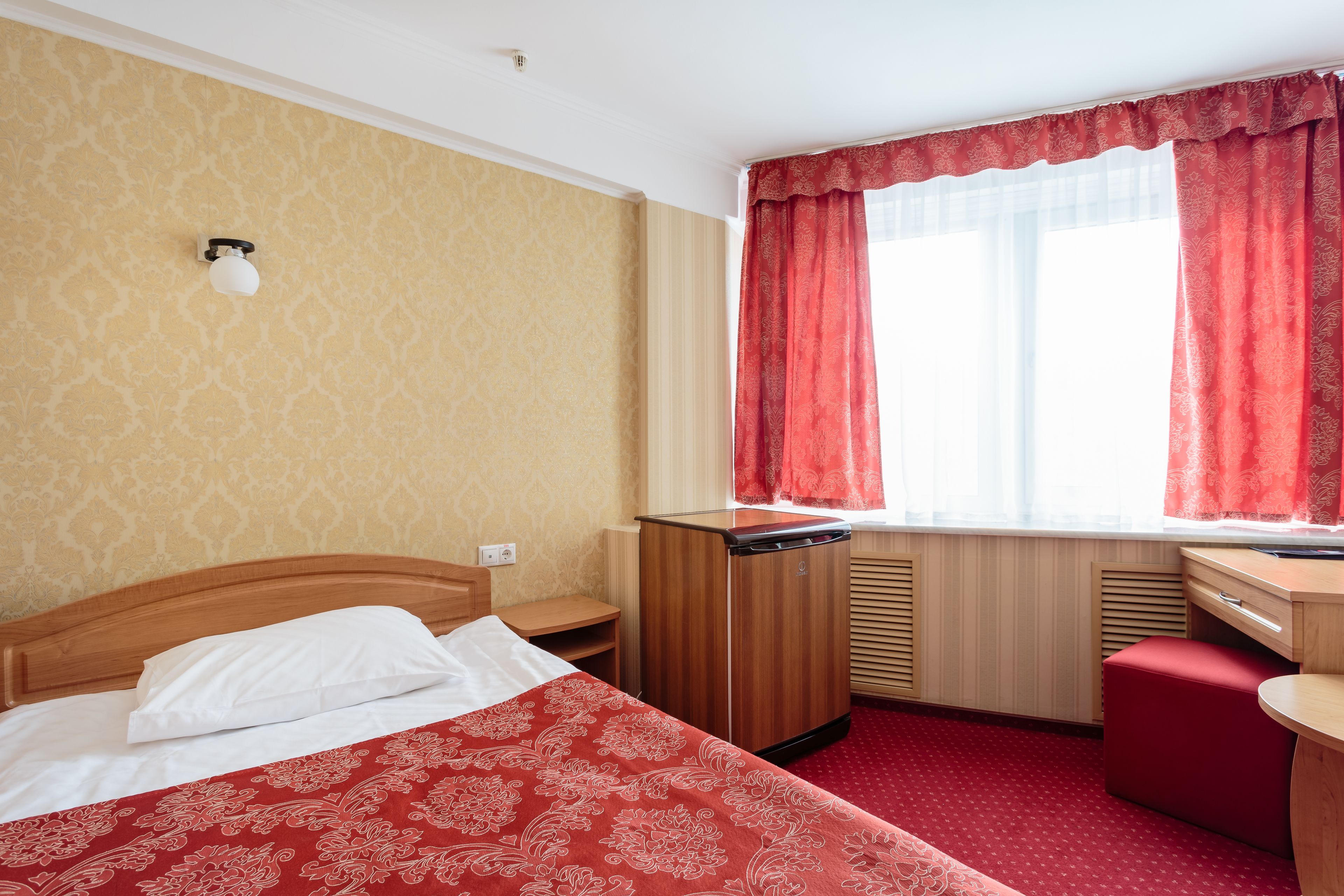 Una habitació d'hotel amb llit, escriptori, radiador i finestra amb cortines vermelles.