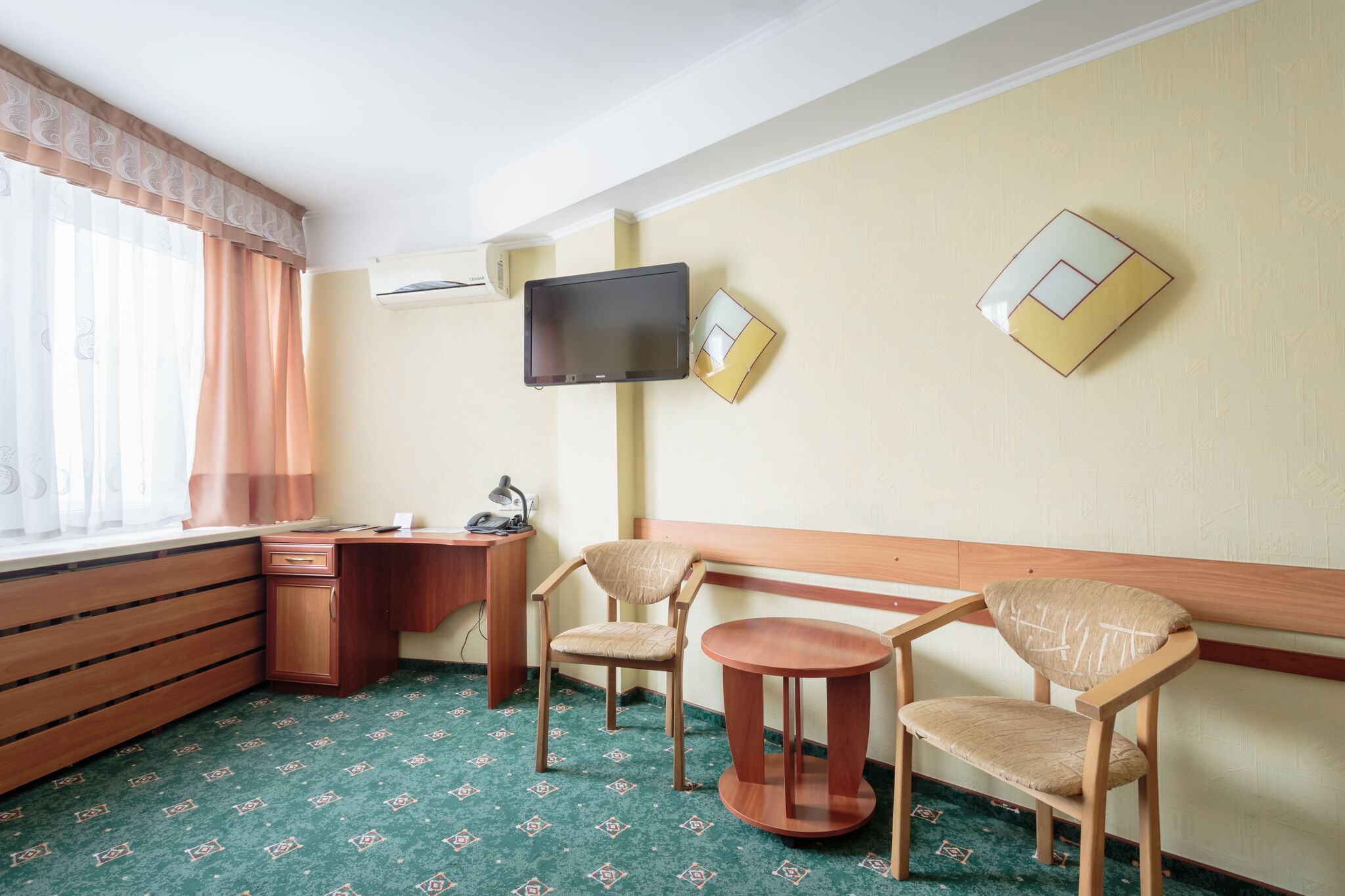 Ett hotellrum med ett skrivbord i trä, två stolar, ett litet runt bord, en tv som är monterad på väggen och ett fönster med gardiner.