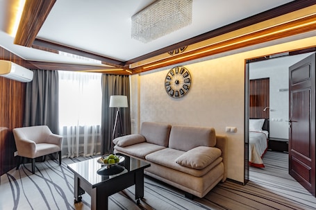 A modern hotel room with a sofa, armchair, and a coffee table with fruit. A large wall clock and a mirror are visible.