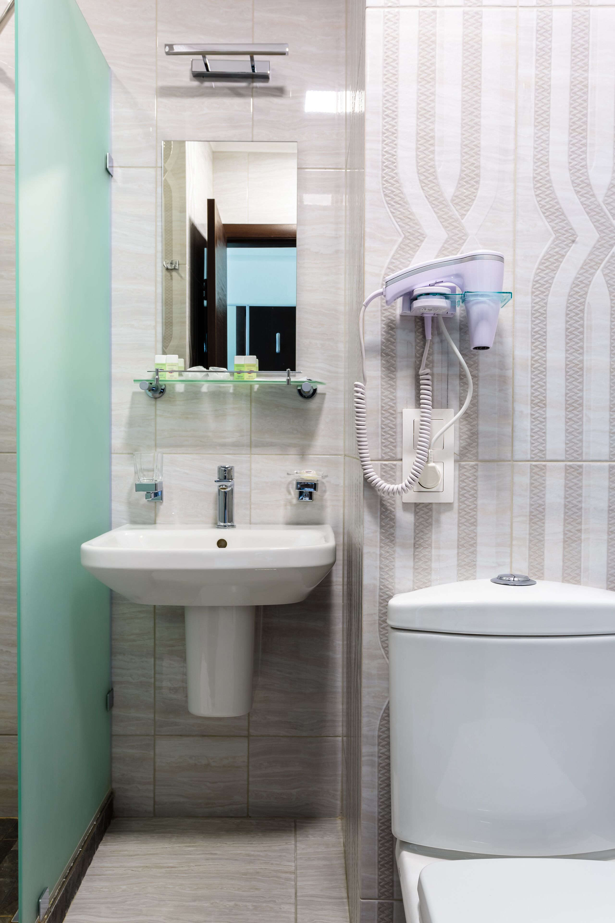 Une salle de bain avec un lavabo, un miroir et des toilettes blancs. Un sèche-cheveux est fixé au mur et une étagère en verre se trouve au-dessus du lavabo.