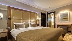 A hotel room with a large bed, a wooden headboard, a nightstand, a desk, and a bathroom visible through an open door. - Premier Hotel Dnister (Lviv)