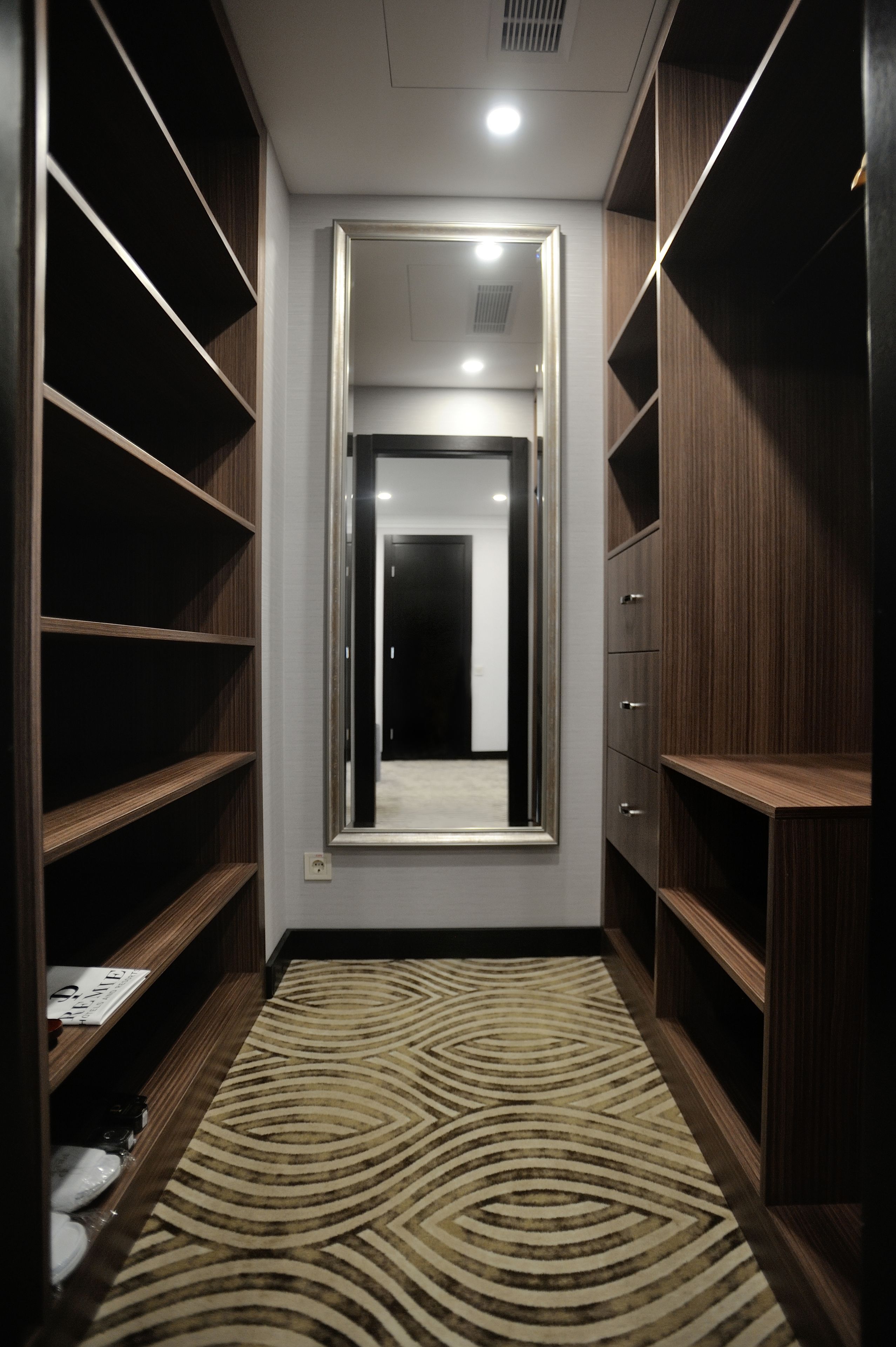 Un pasillo estrecho con estantes de madera a ambos lados, un espejo grande en la pared izquierda y una alfombra estampada.