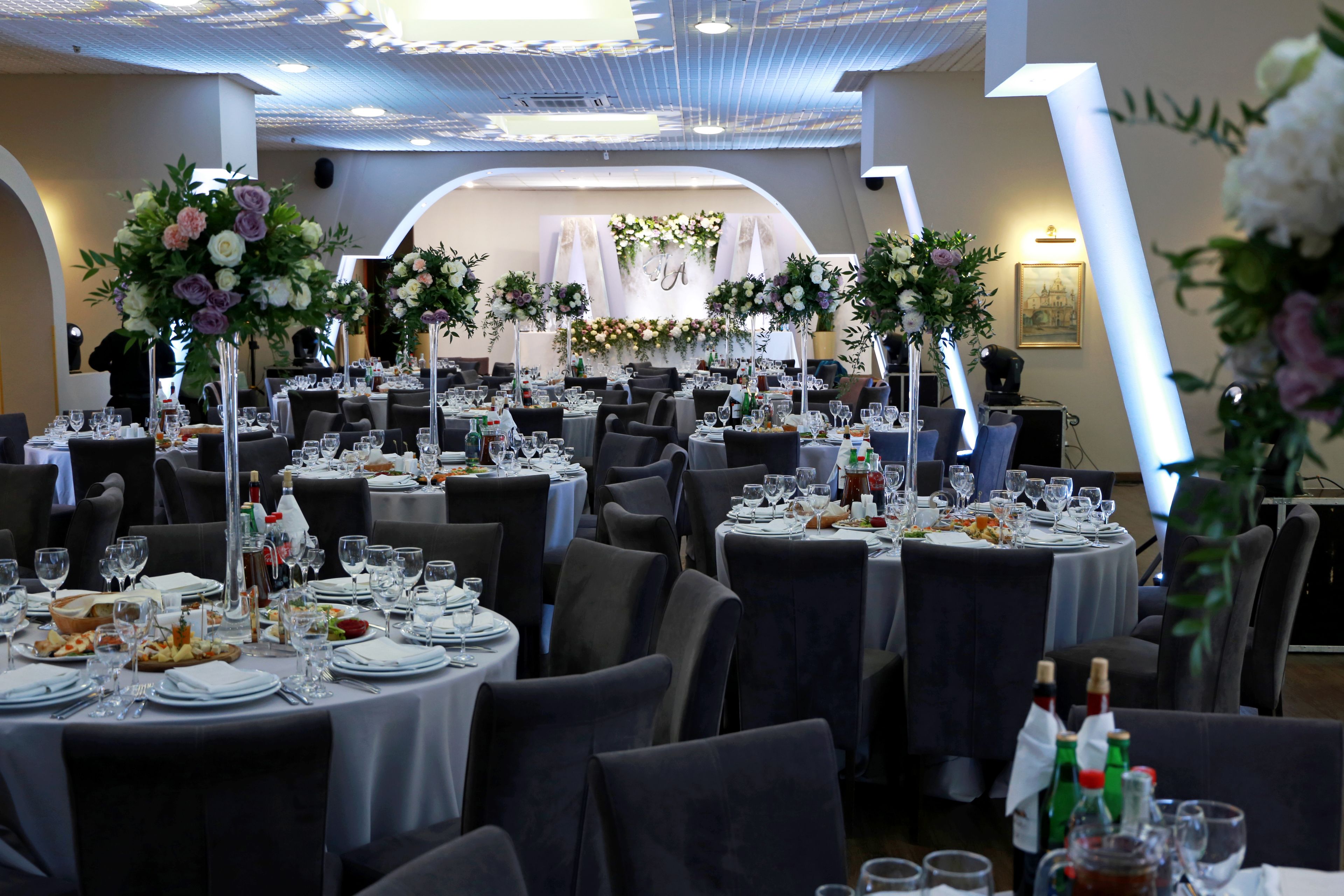 A banquet hall with round tables set for a formal event, featuring floral centerpieces and a chandelier.