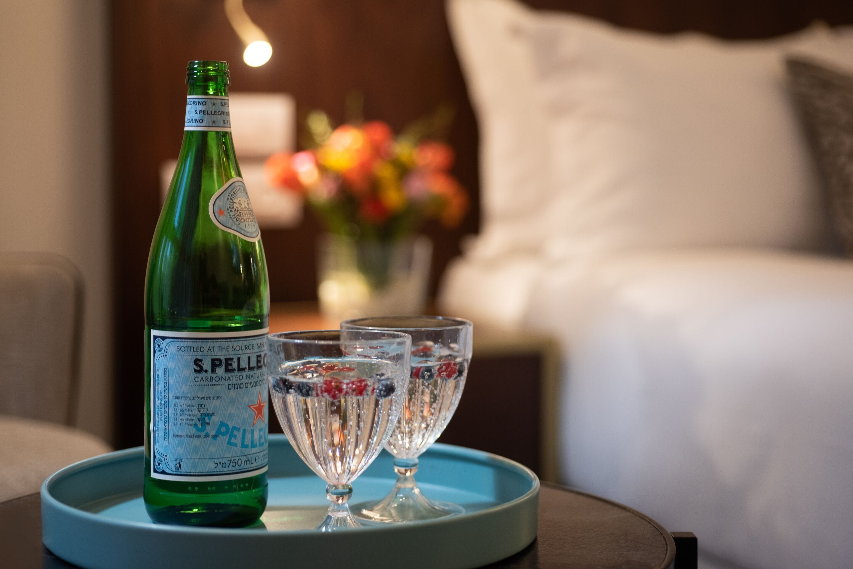 A bottle of S. Pellegrino sparkling water on a tray with two glasses, in a hotel room with a bed and a vase of flowers in the background.