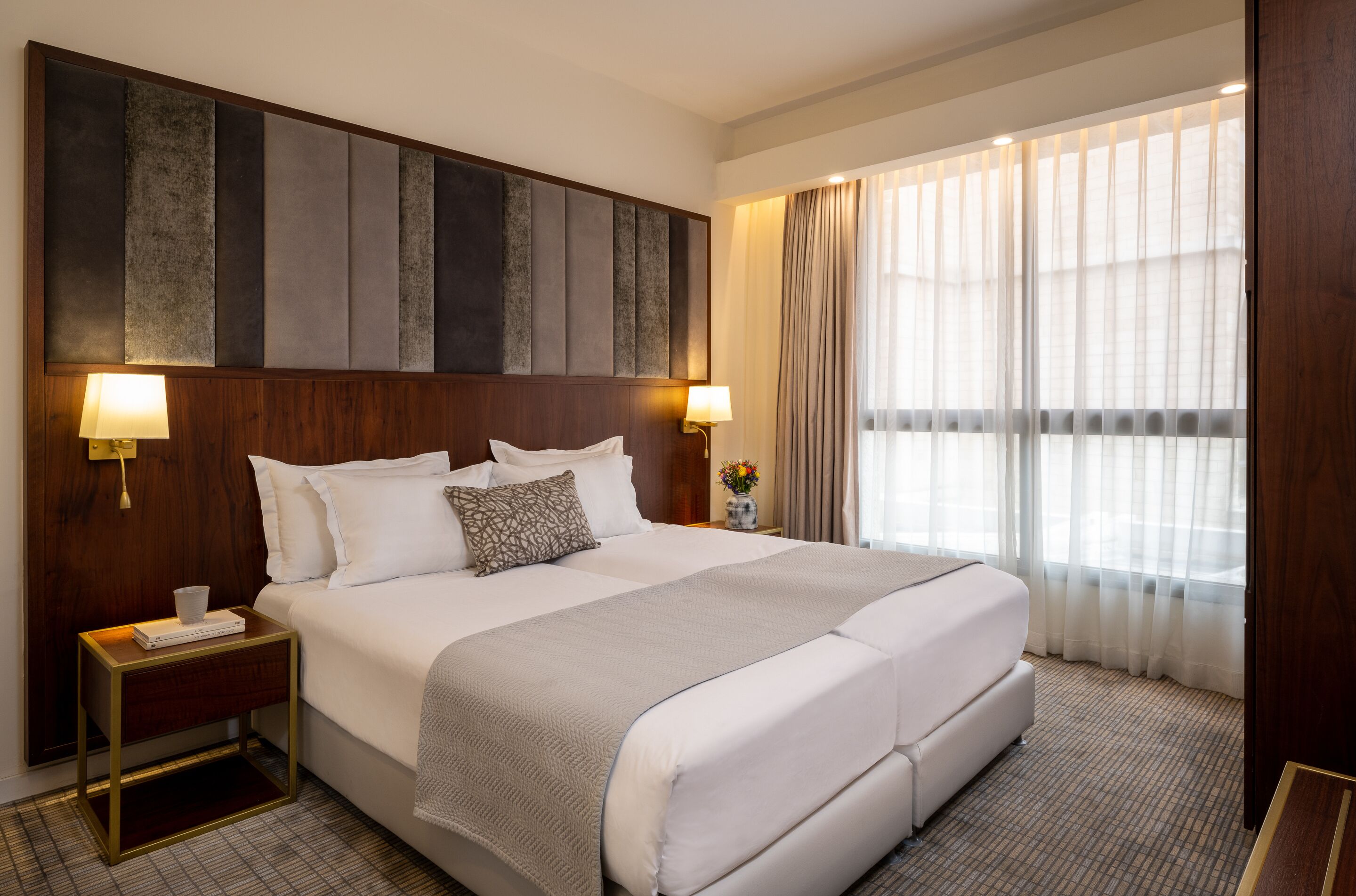 Habitación de hotel con una cama grande, dos mesitas de noche con lámparas, cabecera con panel texturizado y un ventanal con cortinas traslúcidas.
