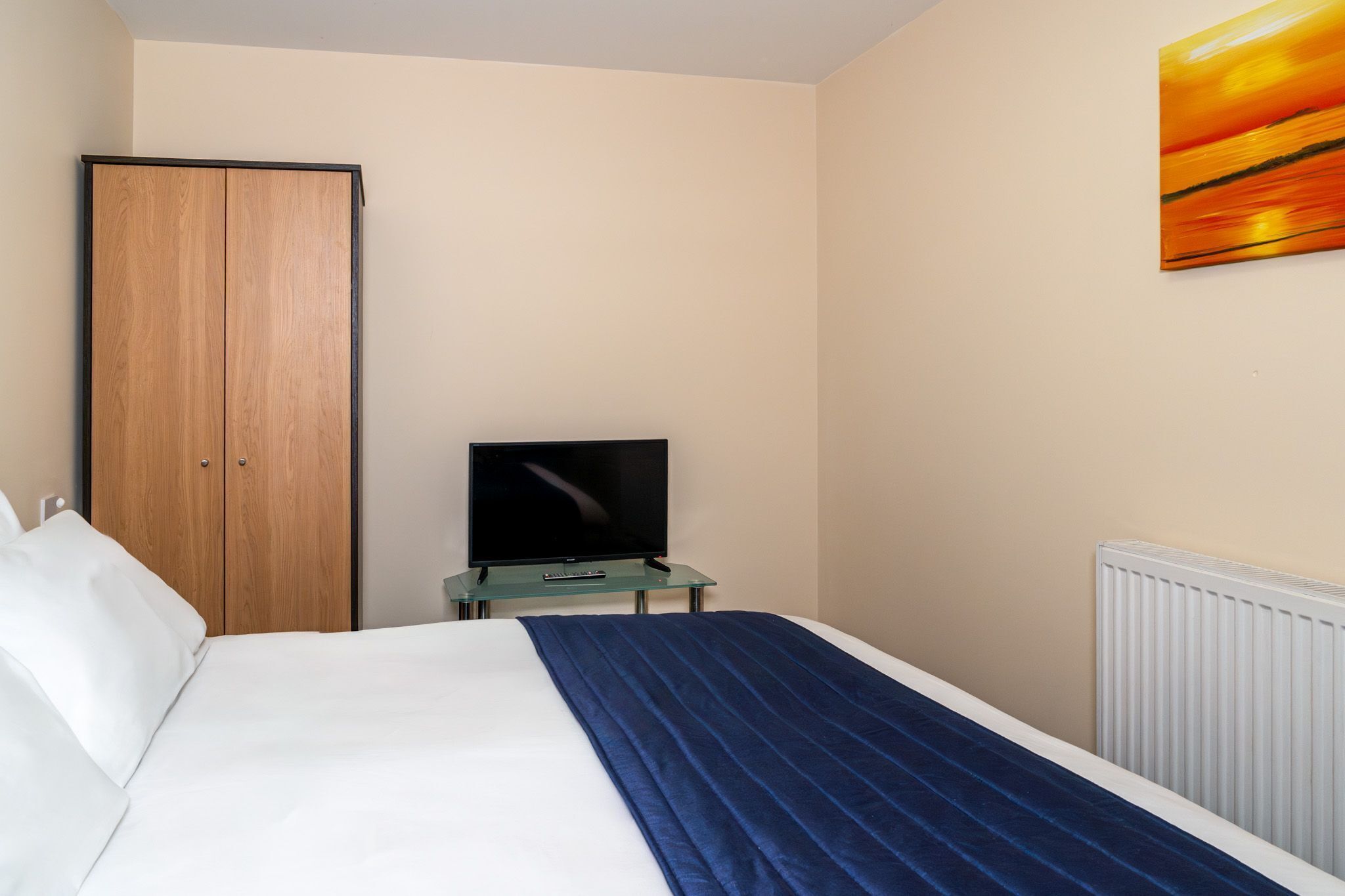 A hotel room with a bed, a television on a glass table, a wardrobe, and a painting on the wall.