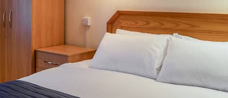 A neatly made bed with white linens, a wooden headboard, a bedside table with a drawer, and a wall socket.