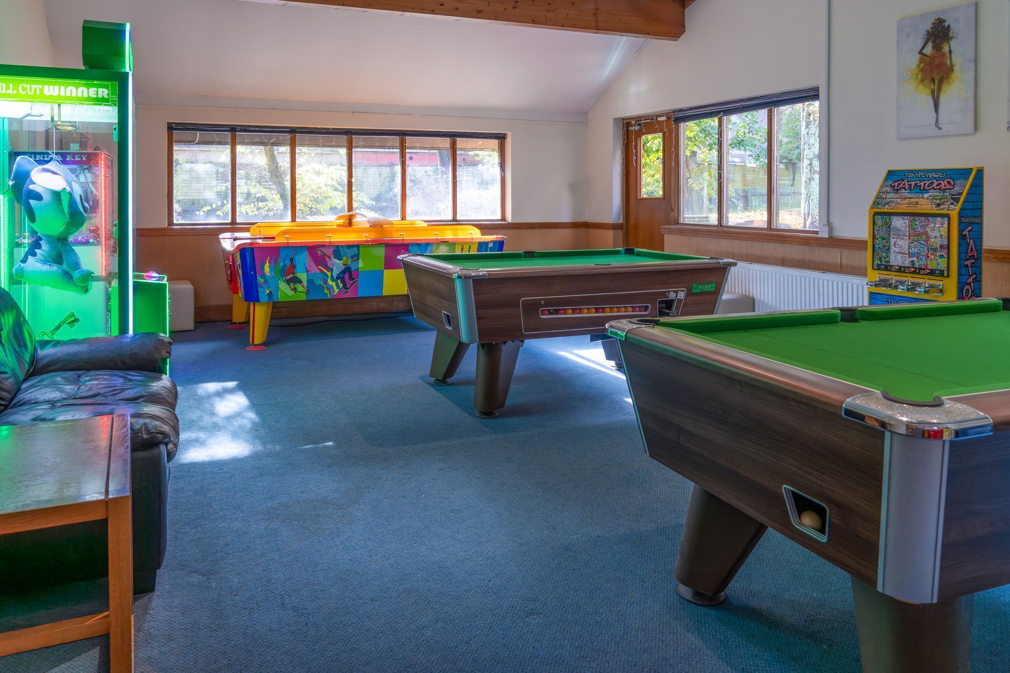 Uma mesa de bilhar, uma mesa de ténis de mesa colorida e uma máquina de garra num espaço com grandes janelas.