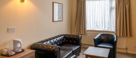 A room with a black leather sofa, a matching armchair, a wooden coffee table, a wall-mounted lamp, a framed picture, and a radiator.