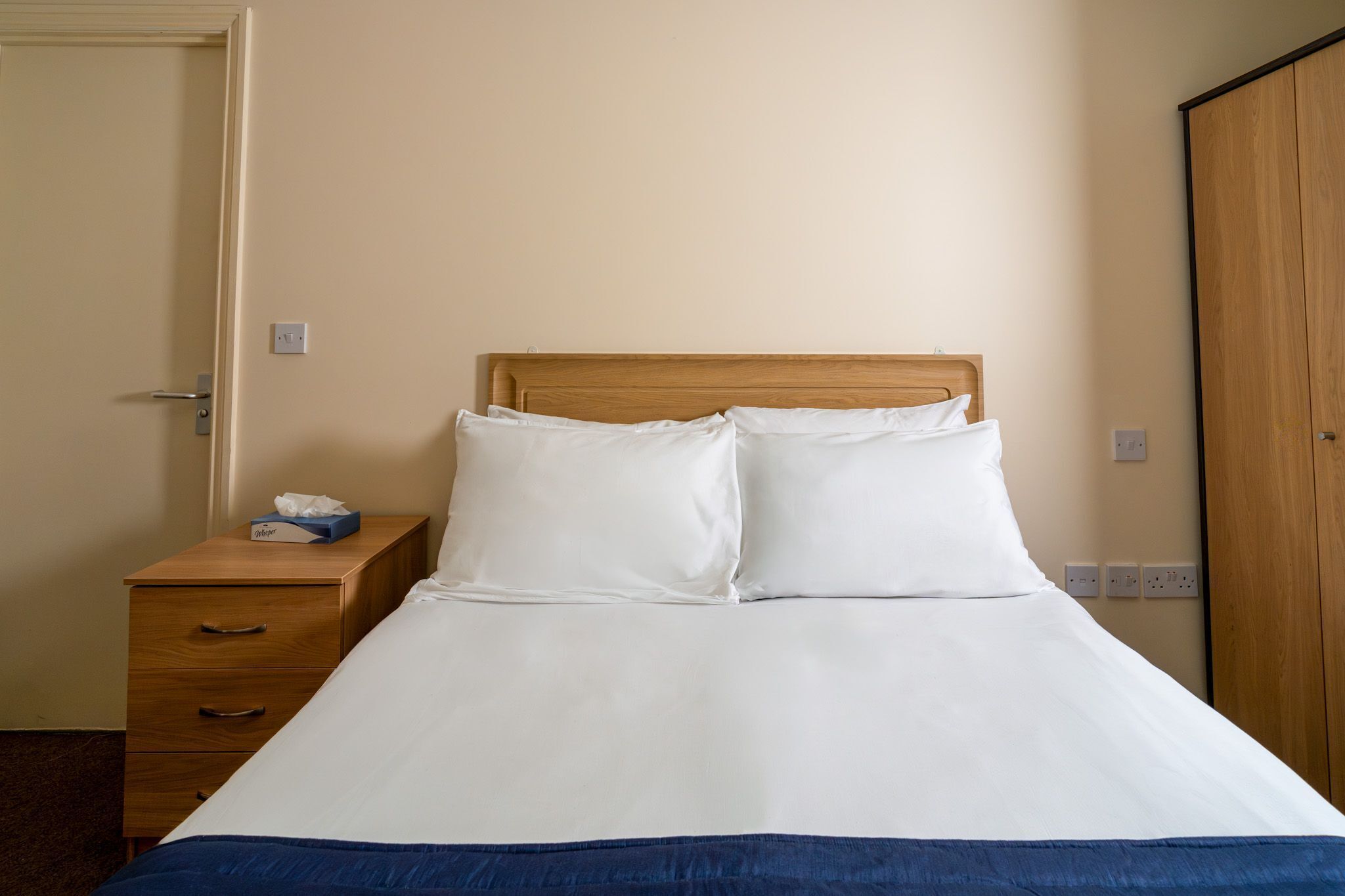 A single bed with white bedding, a wooden nightstand with a tissue box, and a wooden wardrobe.