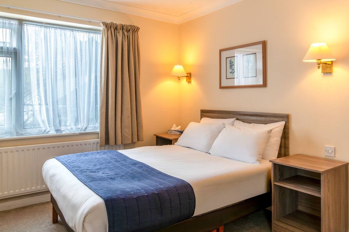 A hotel room with a bed, two bedside lamps, a nightstand, a window with curtains, and a framed picture on the wall.
