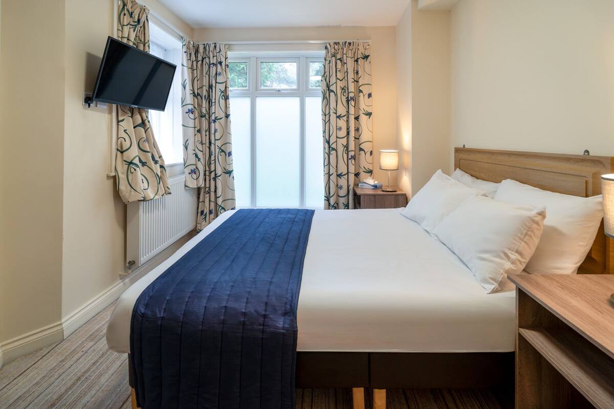 A hotel room with a bed, a TV mounted on the wall, a bedside table with a lamp, and curtains with a floral pattern.